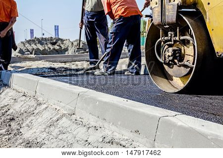 Small steamroller is flatting new road made of asphalt. Detail on newly installed curb. Close up view on roadside.