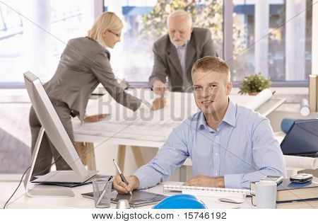 Young confident designer working on drawing pad, looking at camera, smiling, colleagues working in background.?