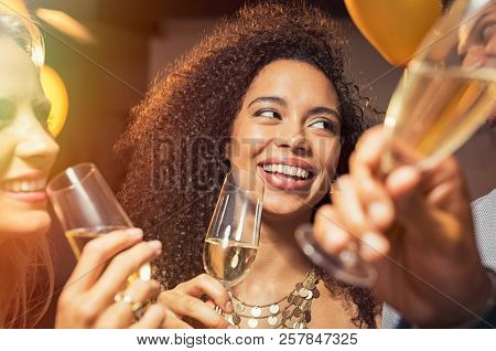 Happy young african woman holding wine glass with friends at party. Portrait of beautiful black girl enjoying new yearâs eve with friends. Cheerful smiling woman drinking champagne at evening party.