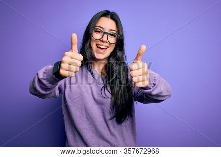 Young brunette woman wearing glasses over purple isolated background approving doing positive gesture with hand, thumbs up smiling and happy for success. Winner gesture.