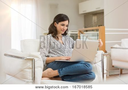 Young woman doing research work for her business. Smiling woman sitting on sofa relaxing while browsing online shopping website. Happy girl browsing through the internet during free time at home.