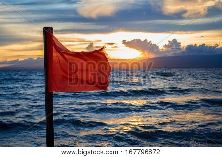 Red flag by the sea with sunset background. Evening seaside scene with danger warning sign. Beach warning flag splashing on wind. Stormy sea threat warning. Vacation danger. Extreme seawater caution