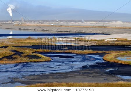 Alternating stripes of bitumen, water, sand and grass at a mine's tailings pond, where fine waste gradually settles. Also one oil barrel visible.