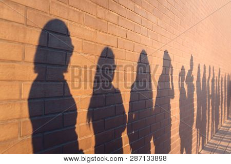 A Line Of Shadows Of People Lined Up Against A Red Brick Wall. Stand In A Queue To The Changes