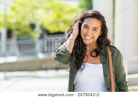 Happy young beautiful woman walking on the street. Portrait of cheerful university student looking at camera while adjusting curly hair with copy space. Latin stylish girl smiling while standing.