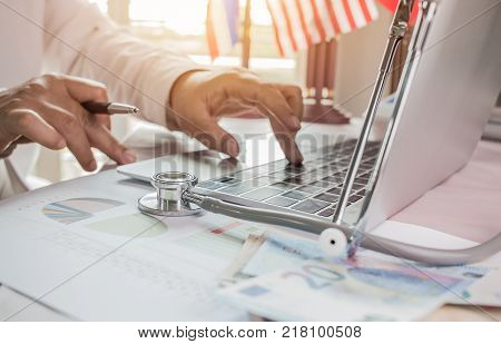 Doctor working on laptop computer with report analysis and money about Healthcare costs and fees in medical hostpital office. Focus stethoscope on table. Healthcare budget and business concept