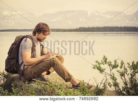 Young Man Traveler with backpack reading book and writing notes outdoor