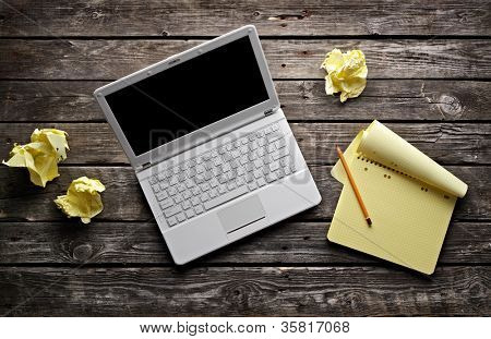 Laptop with blank notepad and pencil with sheets of crumpled paper on old wooden table. Workplace writer or designer.