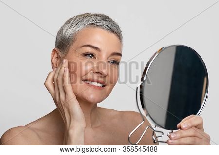 Skincare For Middle-aged Women. Beautiful Mature Lady Looking At Mirror And Applying Anti-aging Crea