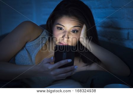 young beautiful Latin woman on bed late at night texting using mobile phone sleepy and tired in internet communication overuse concept and smart phone addiction