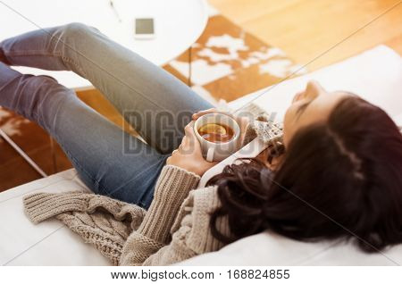 Young woman relaxing on sofa holding cup with lemon tea. Woman dreaming in living room lying on couch with legs on table and looking up. Woman thinking while drinking lemon tea at home.
