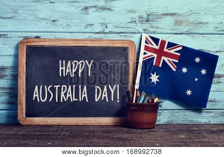 a chalkboard with the text happy Australia Day written in it, a pot with pencils and the flag of Australia, on a wooden surface, against a rustic blue wooden background