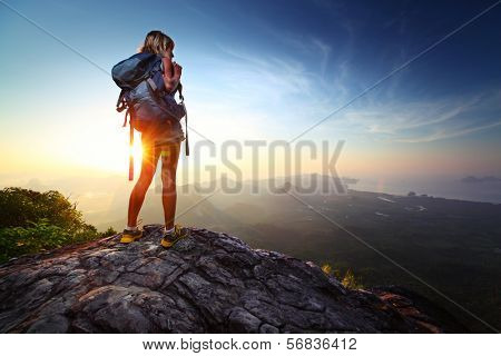 Young lady hiker standing with backpack on top of a mountain and enjoying sunrise