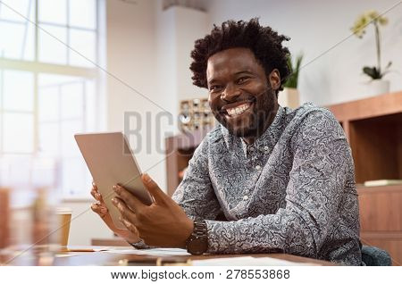 Happy casual african man using digital tablet at office. Portrait of smiling black businessman sitting on chair using tablet pc. African american casual business man looking at camera.