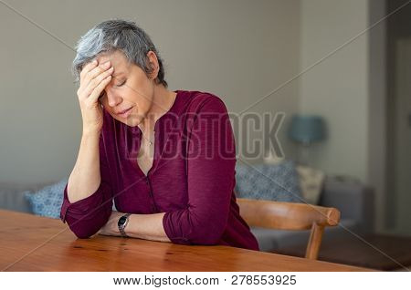 Senior woman suffering from headache while sitting at table in a living room. Depressed mature woman with head in hand thinking. Stressed old lady suffering from migraine at home.