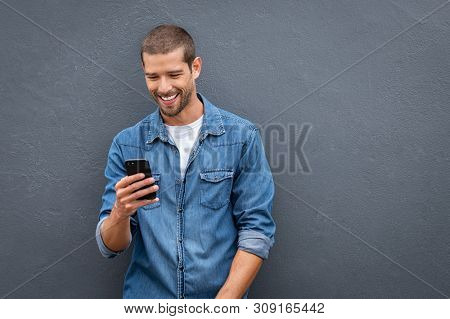 Young man leaning against a grey wall using mobile phone with copy space. Happy casual guy messaging on smartphone on gray background. Cheerful man typing and reading a message on cell phone.