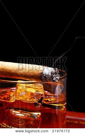 Lit Cigar resting on Glass of Whiskey and Ice cubes with wisp of smoke and black background