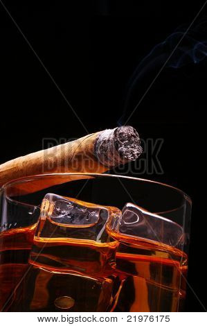 Lit Cigar resting on Glass of Whiskey and Ice cubes with wisp of smoke and black background, low angle