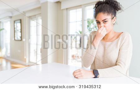 Beautiful young african american woman with afro hair sitting on table at home smelling something stinky and disgusting, intolerable smell, holding breath with fingers on nose. Bad smells concept.