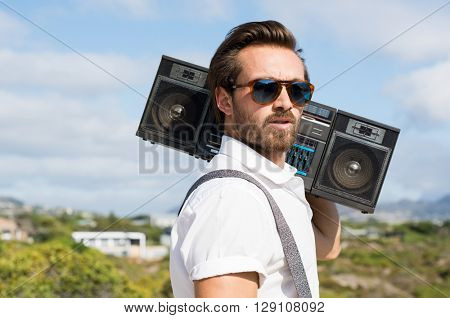 Portrait of a handsome young man holding radio near ears. Close up of hipster guy listening to music on a summer day. Man wearing sunglasses and holding on his shoulder a vintage radio.