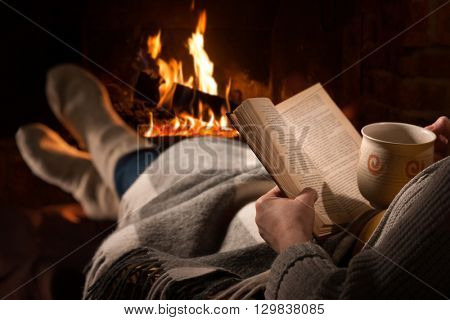 Woman resting with cup of hot drink and book near fireplace