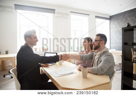 Happy Millennial Couple And Smiling Financial Advisor, Lawyer Or Realtor Shaking Hands At Office Mee