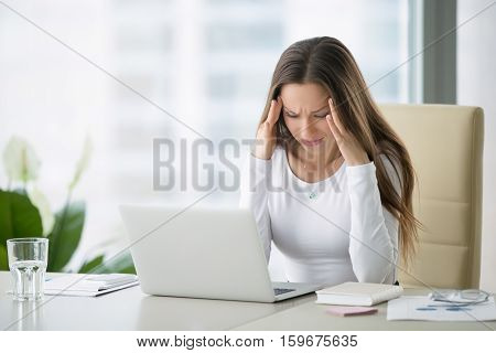 Young frustrated woman working at office desk in front of laptop suffering from chronic daily headaches, treatment online, appointing to a medical consultation, electromagnetic radiation, sick pay