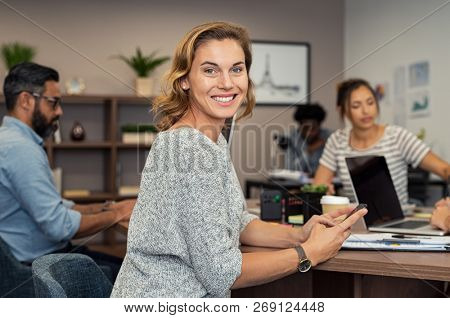 Mature businesswoman using smartphone during business meeting. Successful business woman checking mails and replying from mobile while looking at camera. Portrait of smiling casual lady at office.