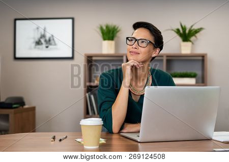 Thoughtful fashion business woman working on a laptop and looking up. Beautiful glamour girl daydreaming at her work station. Latin smiling businesswoman looking up and thinking of business success. 