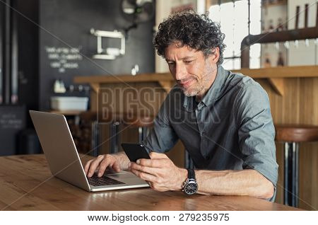 Happy mature business man sitting at cafeteria with laptop and smartphone. Businessman texting on smart phone while sitting in a pub restaurant. Senior man working and checking email on computer.