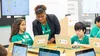 Two students in green shirts sit in front of laptops at a table. A smiling teacher leans over one of the students to also look at the laptop screen.