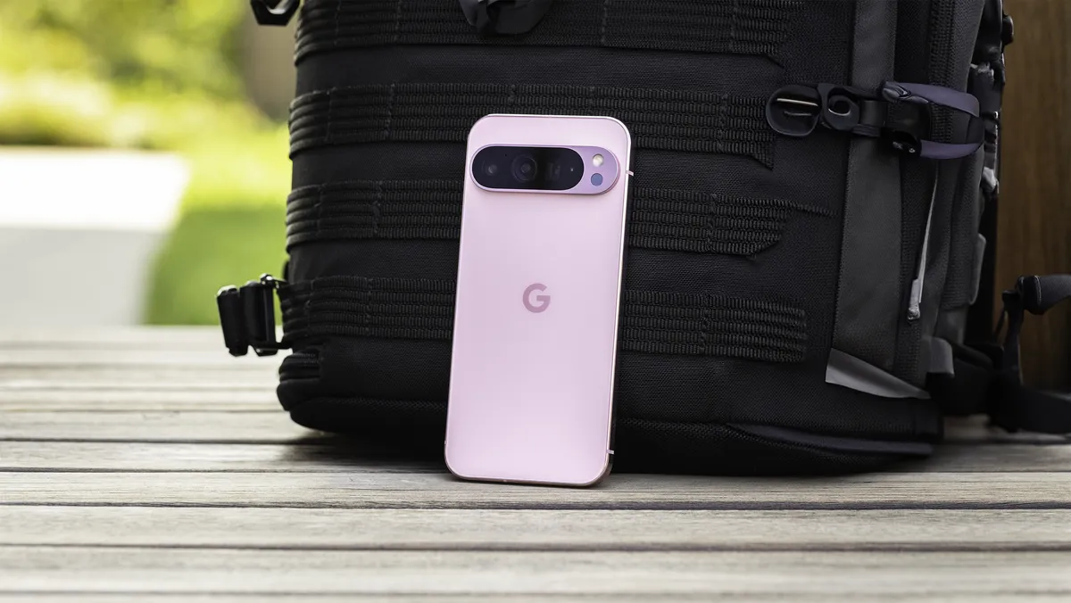 A pink Pixel 9 Pro leaning against a black backpack. The back of the phone is facing the camera, revealing its pill-shaped camera bar and “G” logo in the center.