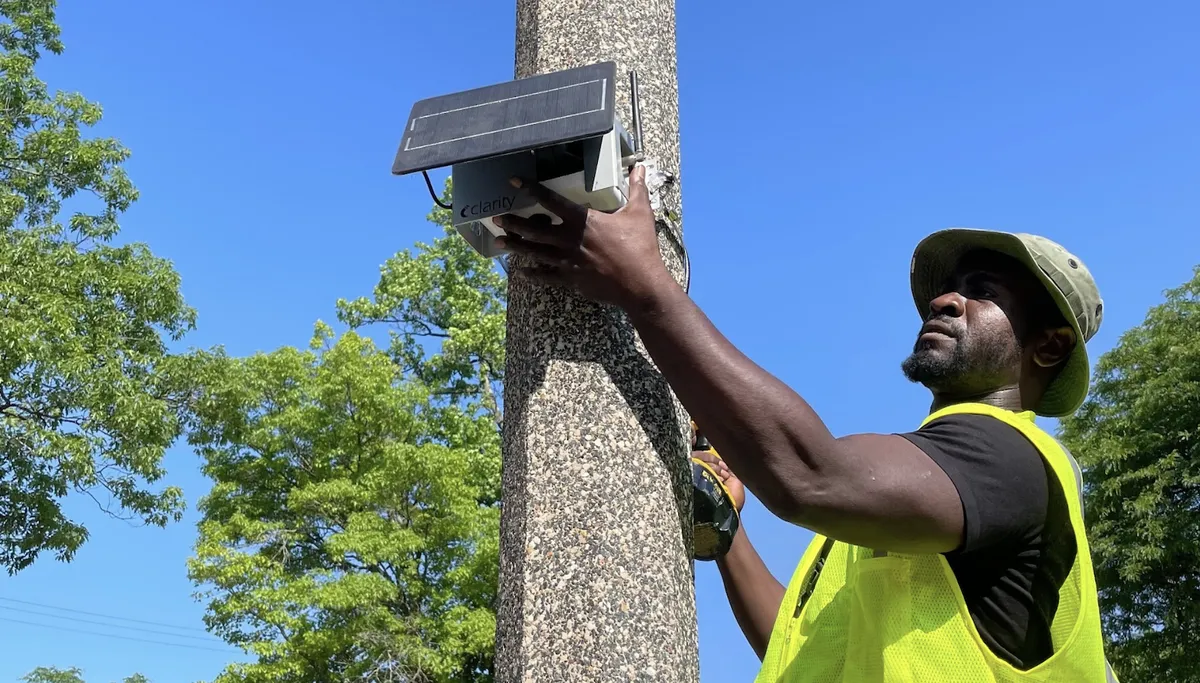 A man wearing a yellow vest hooks up air quality monitoring equipment to a gray lamp post.