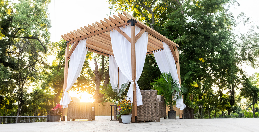 Outdoor wooden pergola with white drapes, surrounded by plants and wicker furniture, set against green trees and sunlight in the background.