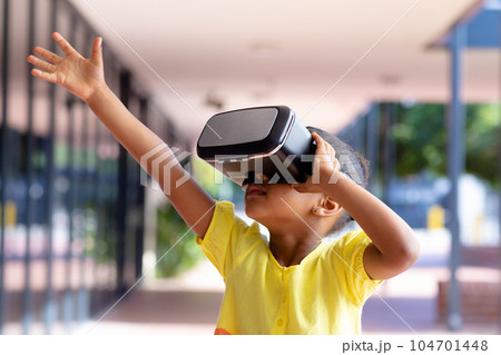 Biracial schoolgirl using vr headset reaching out hand in sunny school corridor with copy space 104701448