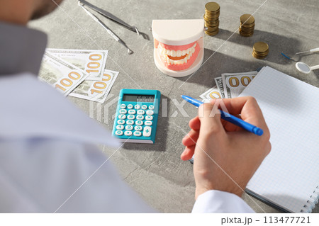 Man with pen in hand, notepad, cash, calculator, jaw and dental instruments on gray background 113477721