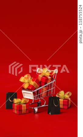 Supermarket trolley with red box with gold bows and black shopping bags. The concept of buying gifts for holidays and sales. Discounts, Black Friday. Red background, Copy space vertical banner 118755524