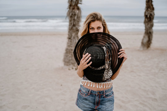 Cheerful blonde woman in colorful top and jean shorts blocking her face with a big hat on seashore