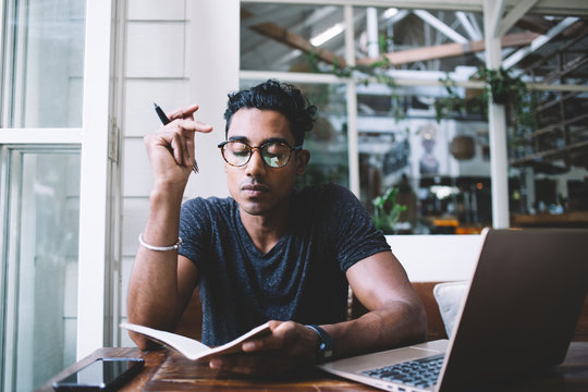 Thoughtful ethnic writer writing in journal while sitting at table with gadgets