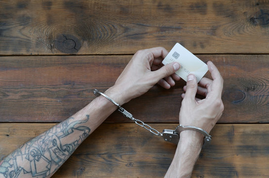 Cuffed hands of tattooed criminal suspect of carding holds fake credit card as evidence for investigation