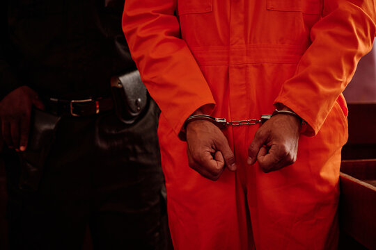 Mid section of young male suspect or prisoner with cuffs on wrists standing next to policeman in uniform bringing criminal to courtroom