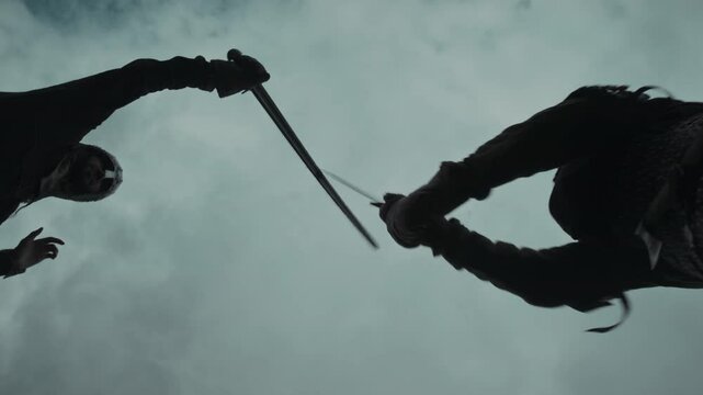 Medium below view, from ground level of two men in medieval warrior armour and chainmail costumes fighting with swords against cloudy sky