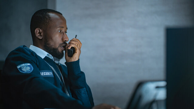 African American security officer or dispatch in uniform uses tablet computer and walkie talkie, looks at screens working in monitoring center during night shift. Surveillance and CCTV system concept.