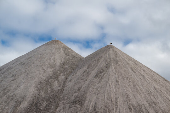 A stockpile of salt or sodium chloride road salt, halite, and rocksalt stored for winter snow and ice deicing controls. The road salt is piled in two mounds with the top portion against a blue sky.