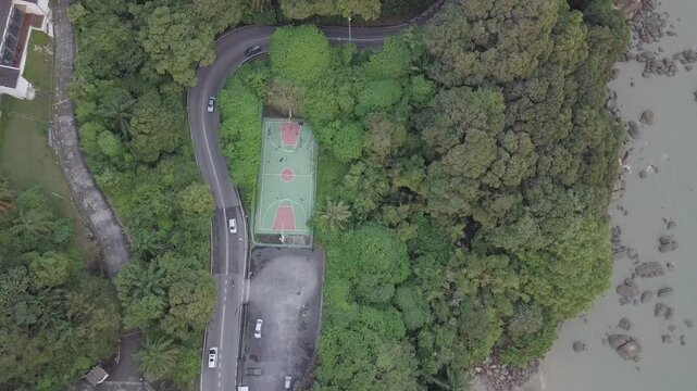 Aerial drone footage of an exotic basketball court by the seaside with a countryside road surrounded by forest. Green nature living. Wellness and wellbeing environment.