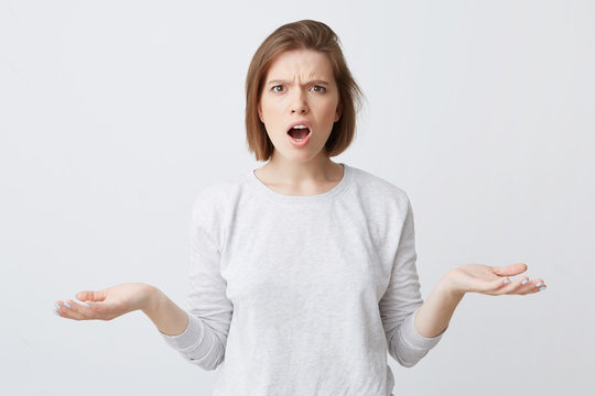 Angry puzzled young woman with opened mouth in longsleeve looking shocked and holding copyspace on both palms isolated over white background