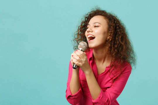 Portrait of beautiful african girl in casual clothes dancing sing song in microphone isolated on blue turquoise wall background in studio. People sincere emotion lifestyle concept. Mock up copy space.