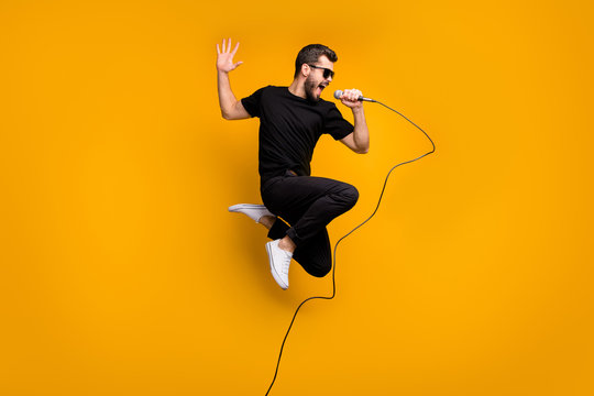 Full body profile photo of crazy hipster guy jumping high holding microphone music lover singing favorite song wear sun specs black t-shirt pants isolated yellow color background