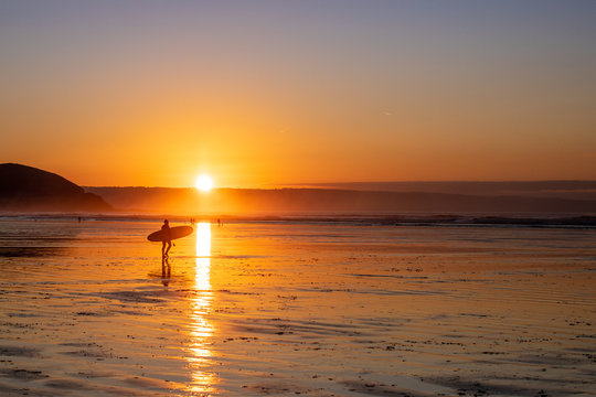 A surfer makes his way down the beach to the waves at sunset on Westward Ho! Beach