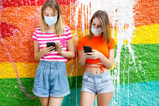 Fashionable female friends in shorts using smart phones against graffiti wall during pandemic